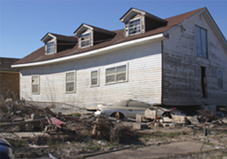 A white colored house with brown roof damaged from Hurricane Katrina with debris surrounding it.