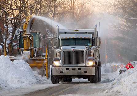 After a winter storm, a snow machine is blowing white snow in to a large, silver dump truck.