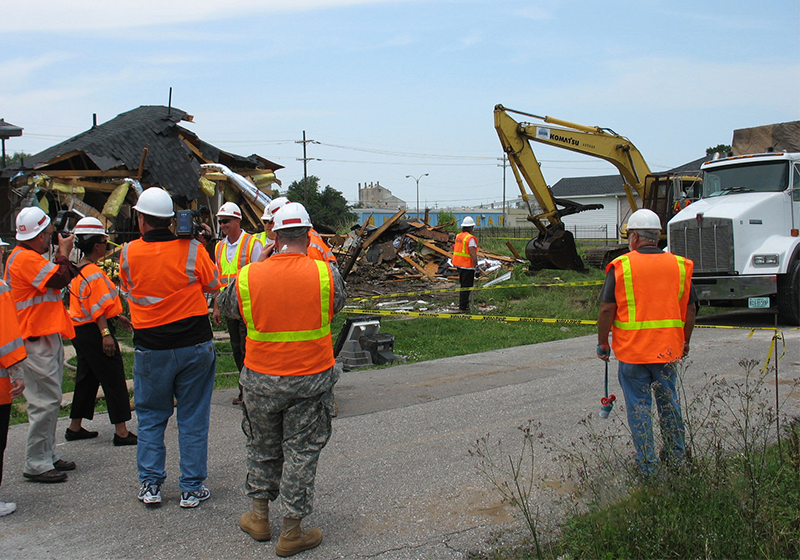 Photo of construction workers working at a house after Hurricane Katrina. Construction workers are in reflective orange vests and white hard hats, observing a vehicle pick up wood debris.